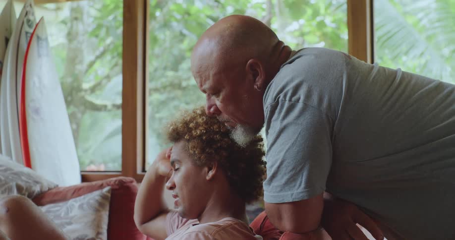 Older man with a white beard leans close to a young man with curly hair who is sitting on a sofa in an indoor setting with surfboards nearby