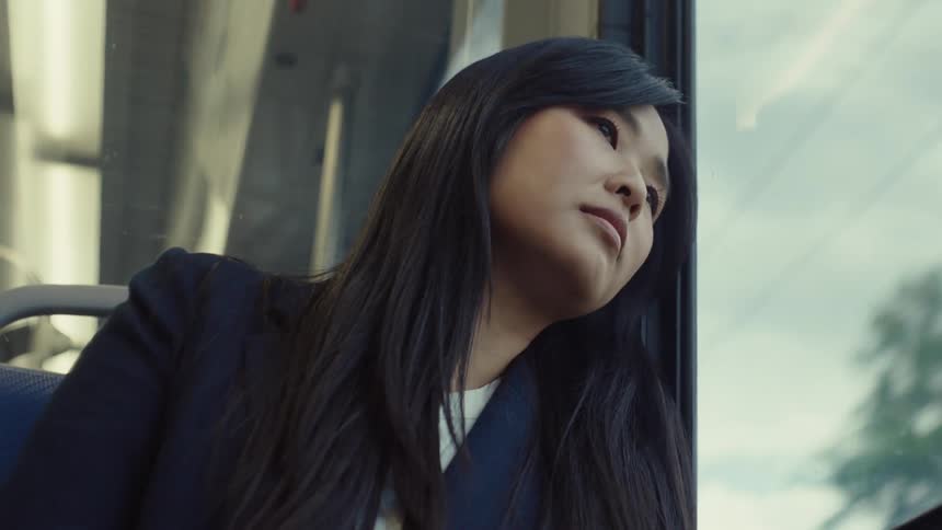 A woman with long dark hair is sitting by a train window resting her head against the glass with a view of the sky outside