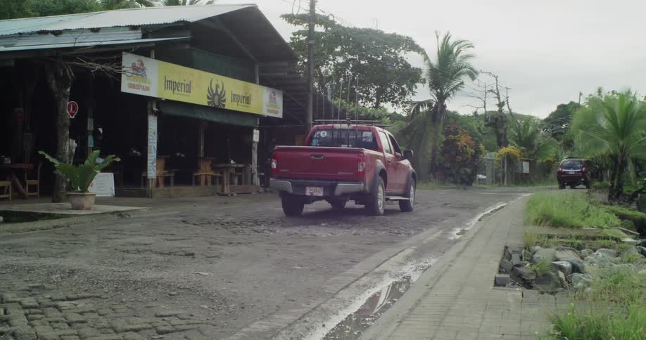 A red pickup truck is parked on a rough cobblestone street in front of a rustic openair restaurant