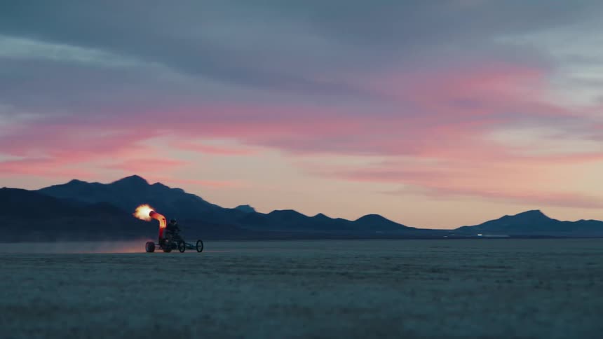 A person rides a homemade vehicle across a vast sandy landscape at sunset propelled by a fiery blast
