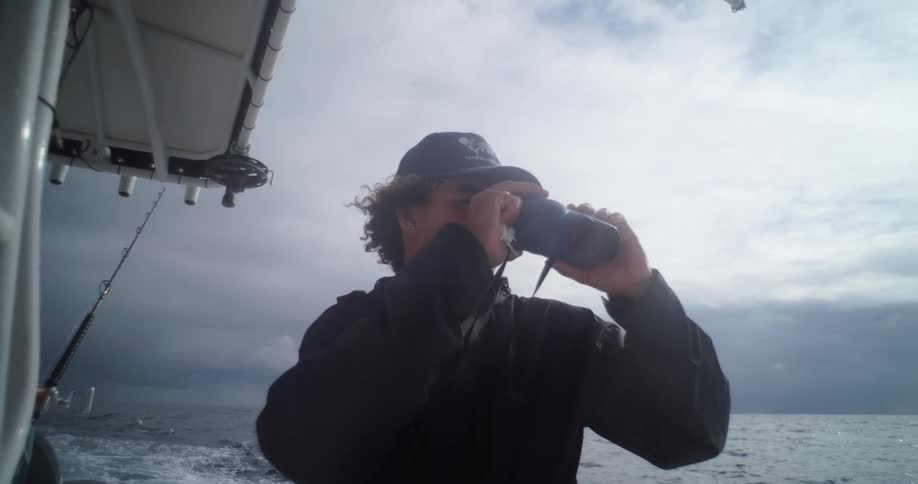 A person on a boat is looking through binoculars at the ocean under a cloudy sky