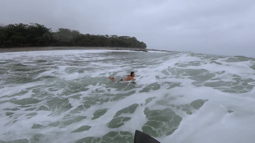 Surfer catches a wave and surfs toward shore while passing another surfer on an overcast day