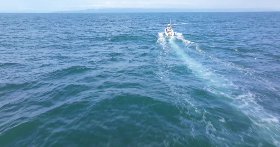 A small white boat is moving through the open ocean leaving a trail of white wake behind it as it heads towards the horizon