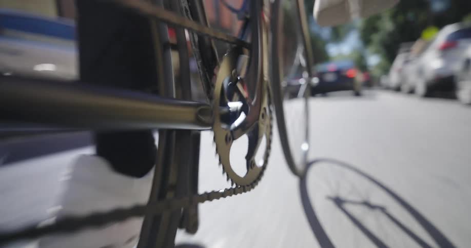 A closeup shot of a bicycles drivetrain focusing on the chainring and pedal with a person wearing white sneakers pedaling on a city street
