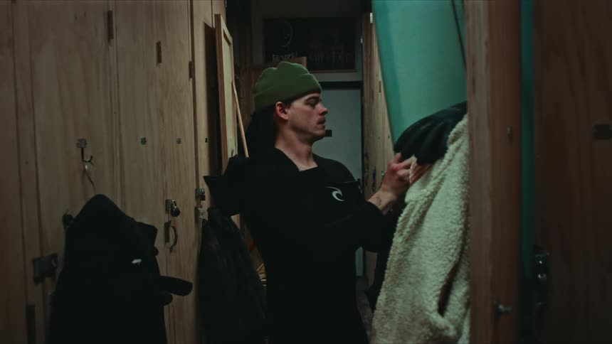 A man wearing a beanie and dark clothing looks up in a locker room then pulls out a surfboard