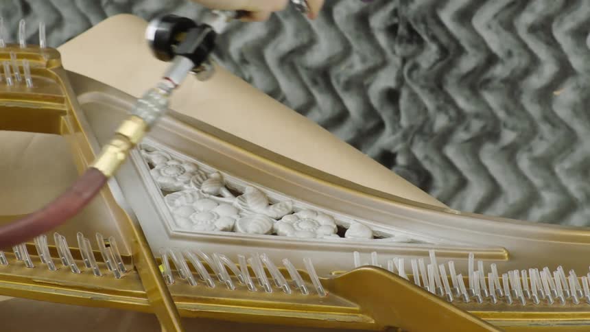 A person uses a spray gun to apply a finish or paint to an ornate wooden section of a grand piano