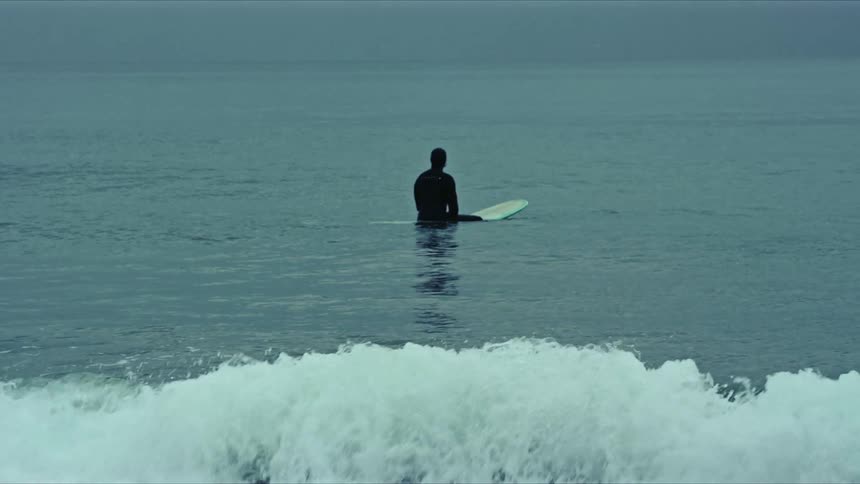 A lone surfer in a wetsuit sits on a surfboard in calm open water under an overcast sky