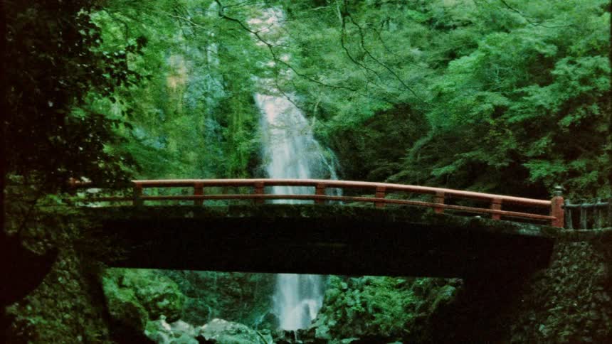 A waterfall cascading behind a traditional red bridge surrounded by lush vibrant green foliage