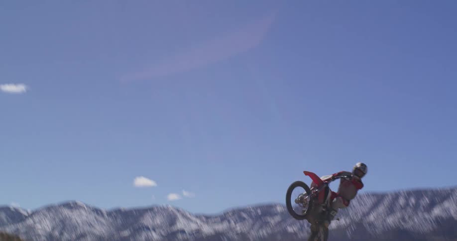 A person is riding a dirt bike on a rugged trail in a mountainous area with snowcapped peaks in the background