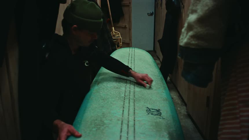 A person is holding and waxing a green surfboard in a narrow dimly lit room with wooden walls and various hanging garments