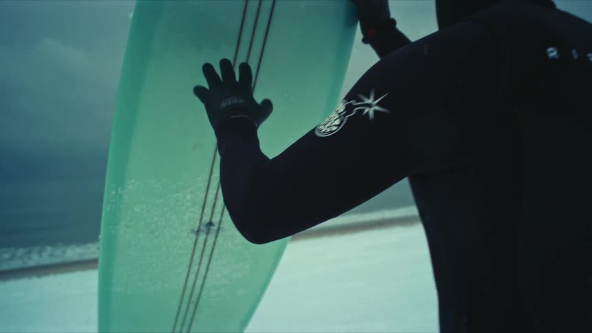 A person in a dark wetsuit and gloves holds a light green surfboard on a beach against a gray sky and ocean