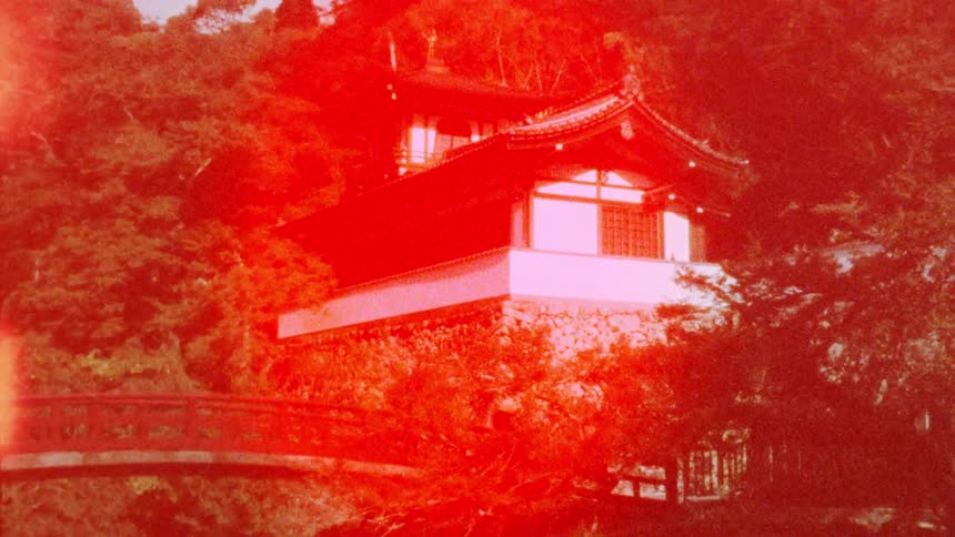 A traditional japanese building with a curved red bridge in the foreground surrounded by dense forest