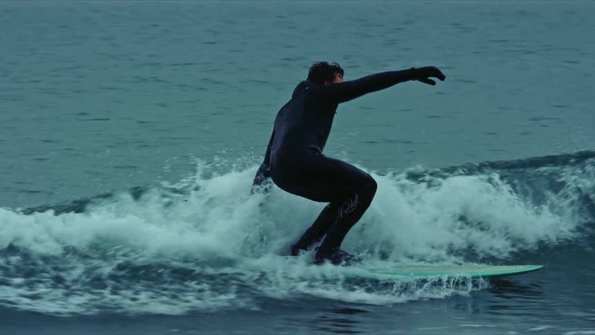 A man wearing a black wetsuit rides a surfboard on a shallow wave