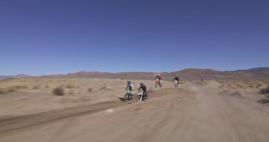 Four motorcyclists are riding offroad in a desert landscape with mountains in the background under a clear blue sky
