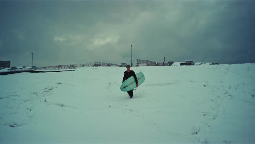 A man carrying a surfboard walks through the snow