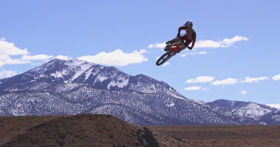 Dirt bike rider jumps his bike between two dirt mounds while snow covers the mountains in the background