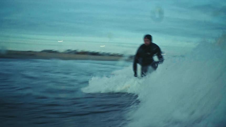 A person is surfing on a wave in the ocean near a coastal city during early morning or late evening with the city skyline visible in the background