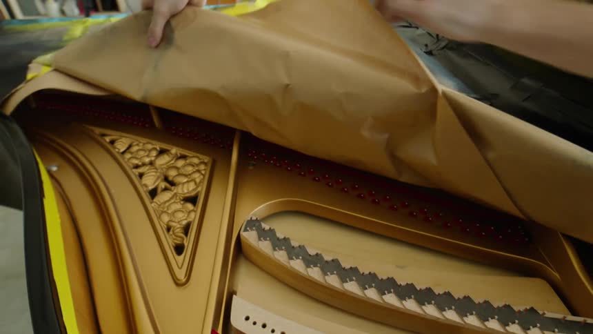 A person is carefully removing brown paper from the interior of a grand piano revealing intricate designs and red accents on the soundboard