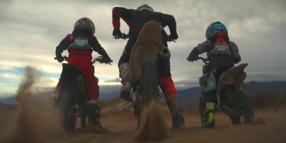 Three people on dirt bikes are seen from behind in a desert landscape then a cloud of dust obscures the view