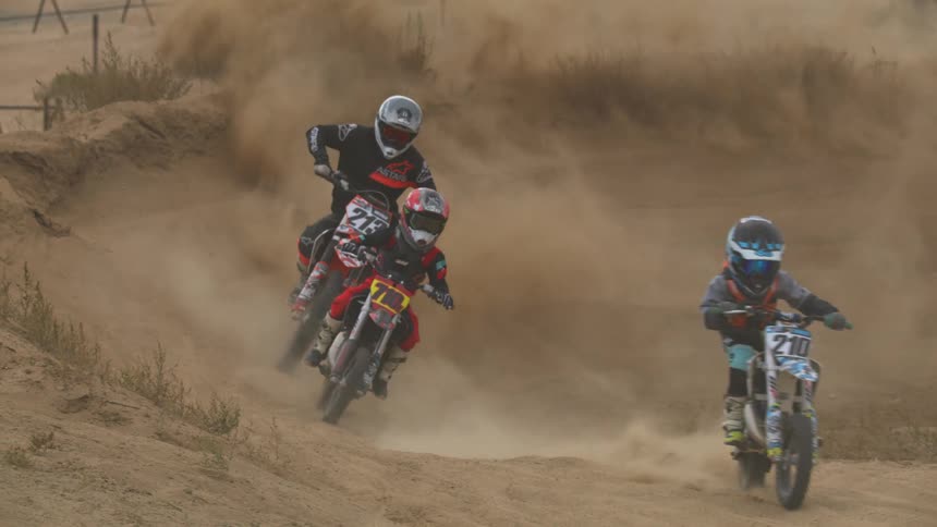 Three dirt bike riders race around a sandy track kicking up dust and vying for the lead