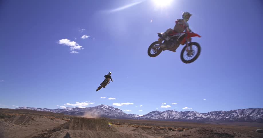 Two dirt bike riders perform jumps against a backdrop of mountains and a bright sky