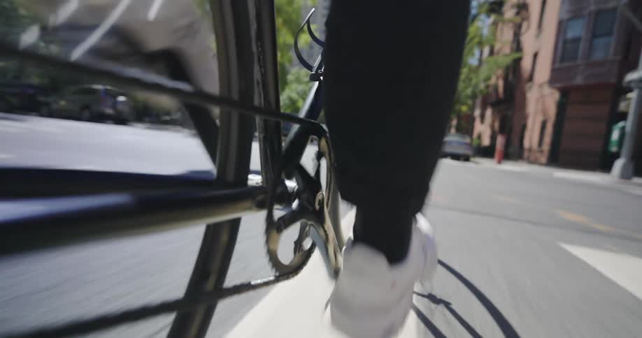 The low angle video follows a cyclists feet pedaling a bicycle down a city street with cars and buildings passing in the background
