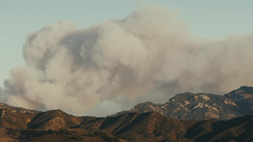 A large plume of smoke rises above a range of rugged brown mountains under a clear sky