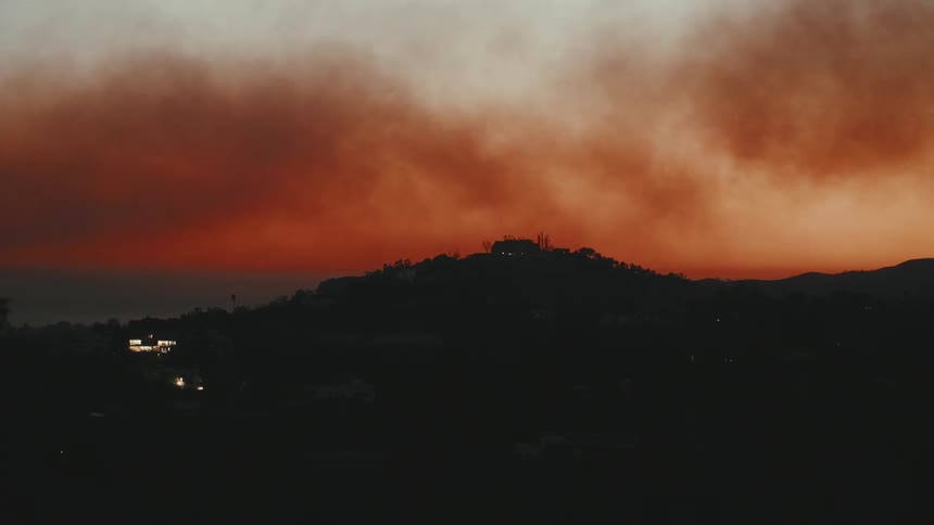 A dark silhouette of a hill set against a dramatic sky filled with thick reddishorange smoke from a fire or intense sunset