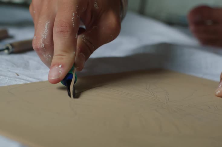 A hand is using a carving tool to engrave a design into a piece of wood or linoleum