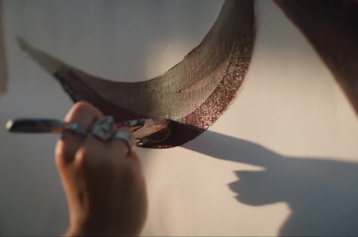 A hand holding a paintbrush is painting a curved dark stroke on a canvas