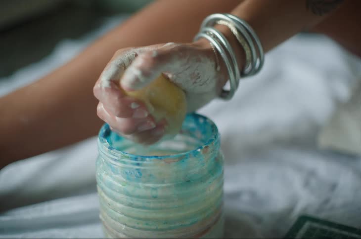 A person with their hands covered in paint holding a sponge and squeezing it into a jar of blue paint