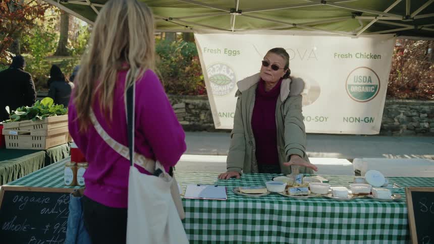 A person in is standing at a market stall with a green and white checkered tablecloth engaging with a vendor pointing at various products on the table