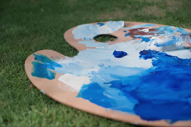 A woman mixes blue and white paint on a palette laying in the grass
