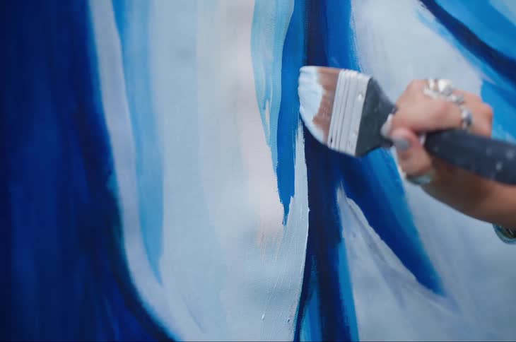 A hand holding a paintbrush is applying blue and white paint in broad strokes on a canvas