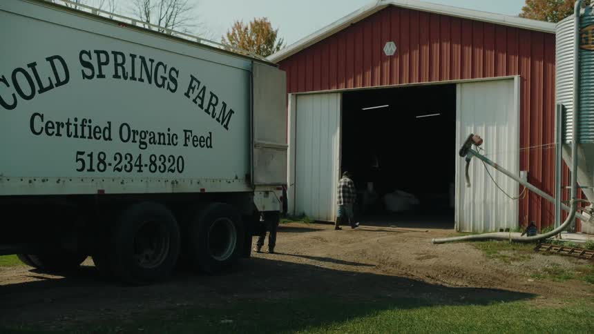 A woman stands at the back of a farm truck as a man goes into the barn