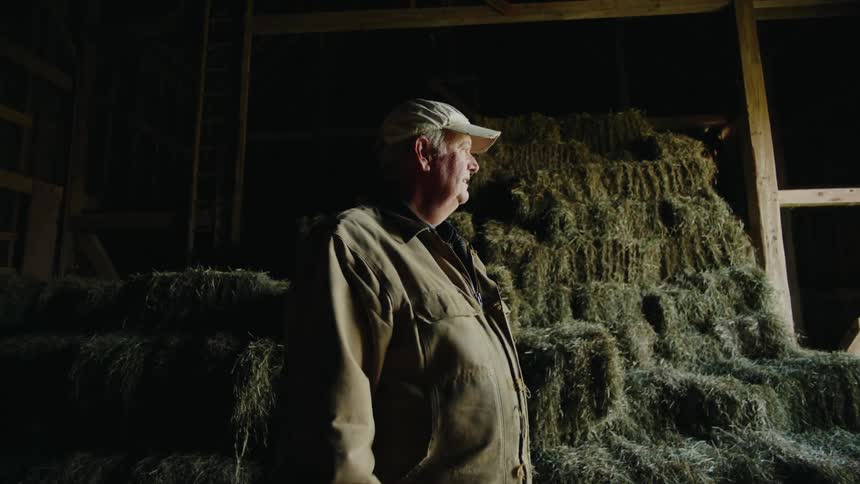 An older man wearing a cap and jacket stands in a dimly lit barn surrounded by stacked bales of hay with light highlighting the rustic atmosphere