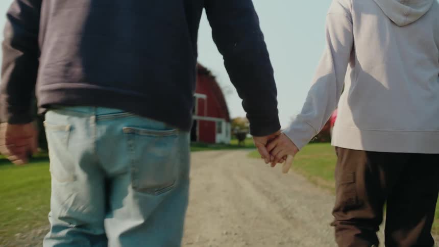 An elderly couple hold hands walking towards a barn on a farm