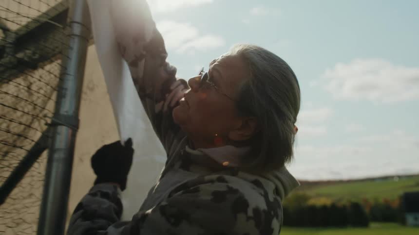 A woman wearing a camouflage jacket and black gloves is adjusting a tarp over a large netted structure in an open field