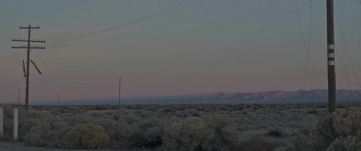 A rural landscape at dusk features multiple utility poles and power lines stretching across the scene