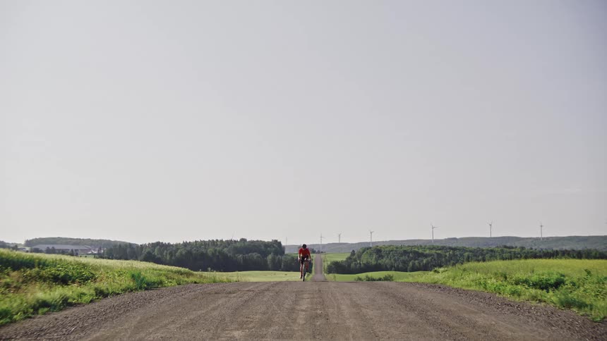 Pass by of a lone cyclist rides along a gravel road surrounded by lush green vegetation and rolling hills in a rural landscape