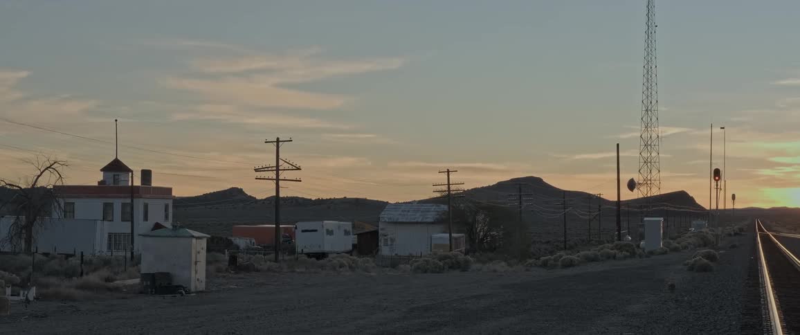 A railroad track stretches towards the horizon as the sun sets next to a small town street lined with power lines and abandoned vehicles