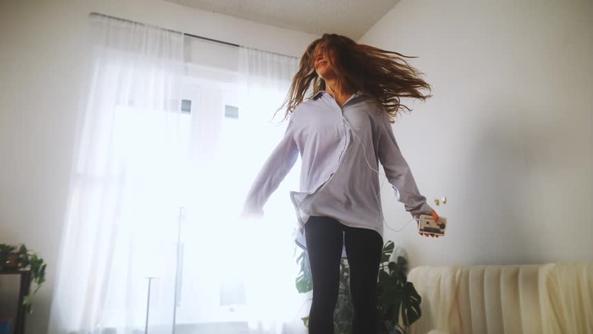 A woman is dancing in a bright room with sheer white curtains holding a phone connected to earphones