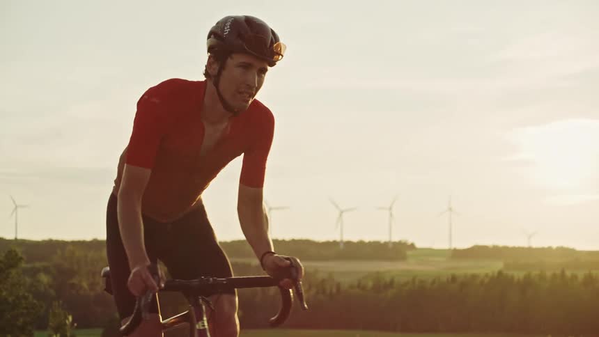 Follow of a male road cyclist at sunset riding along a winding gravel road surrounded by lush green vegetation, wind turbines and rolling hills and in a rural landscape