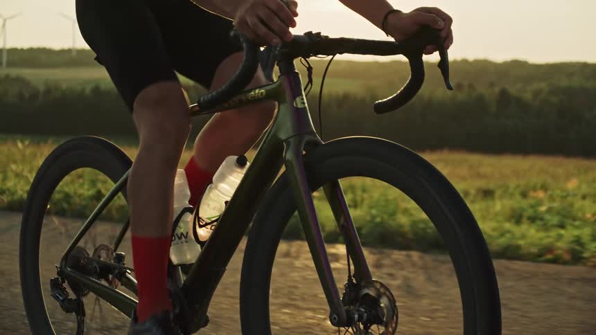 A male road cyclist at sunset riding on a road surrounded by lush green vegetation, wind turbines and rolling hills and in a rural landscape