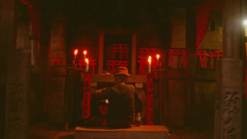 A man sits alone in a dimly lit room in front of a shrine adorned with lit candles