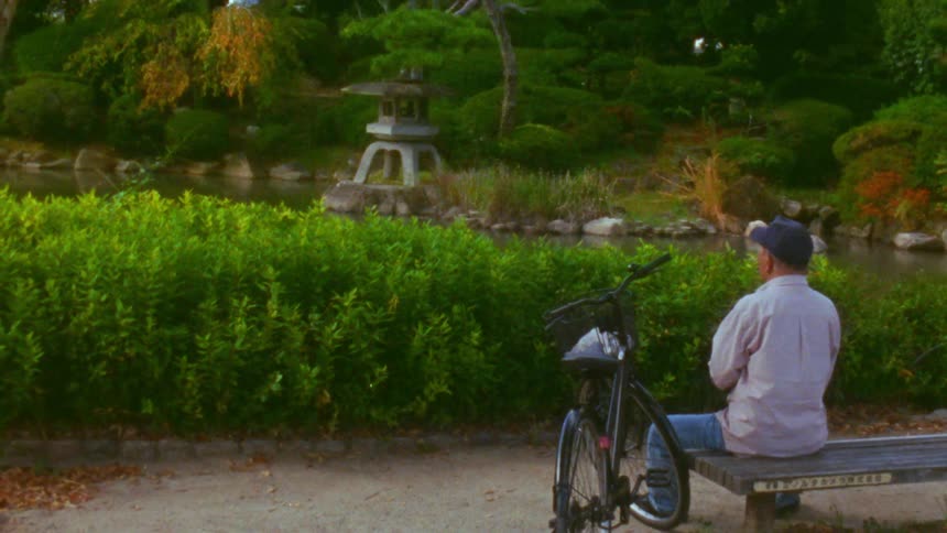 A man is sitting next to his bike in a park looking at a pond