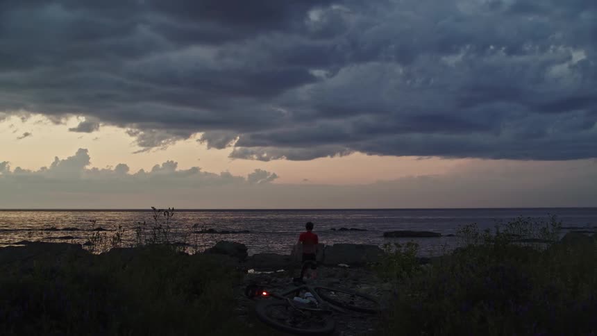 Cyclist arriving at destination in front of a lighthouse at dusk, leaving his bike and walking towards the sea