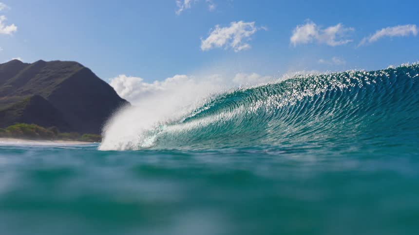 A large curling ocean wave breaking with a mountainous coastline and a clear blue sky in the background
