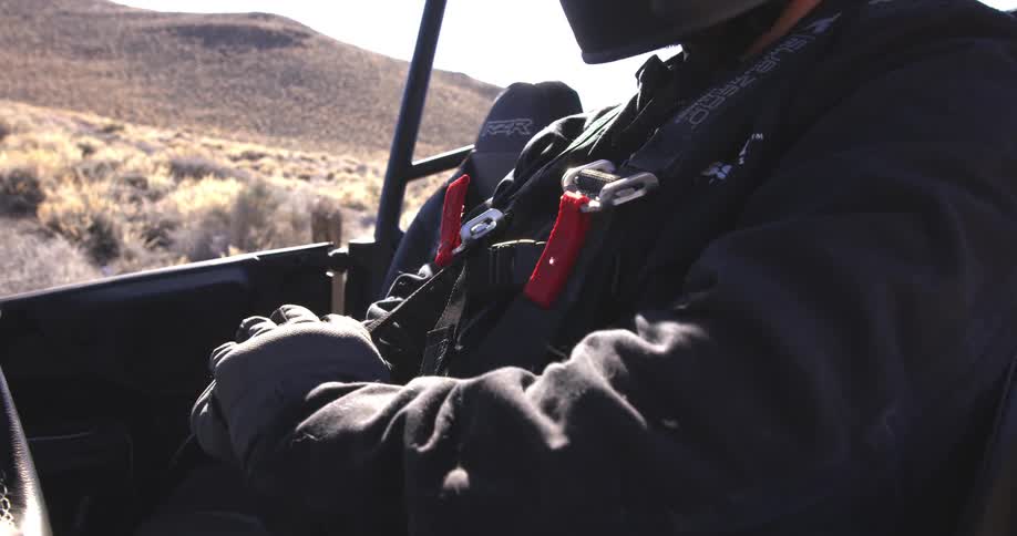 A person wearing a helmet and black clothing is seated in an offroad vehicle securing their harness with a desert landscape visible in the background