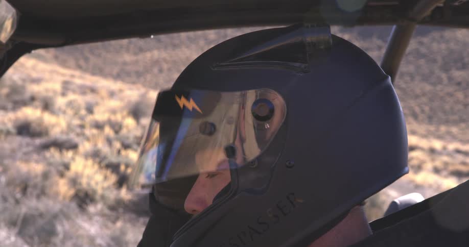 A pilot wearing a black helmet with a clear visor and a yellow lightning bolt symbol sits in a vehicle with a desert landscape visible in the background